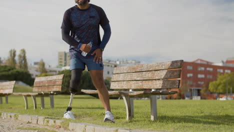 long shot of a tired sportsman with artificial leg drinking water on bench in the park and relaxing after intense cardio workout in the morning