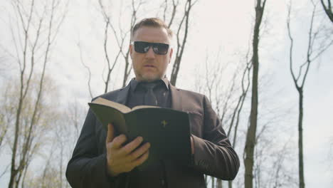 vista inferior de un hombre con traje negro y gafas de sol leyendo una biblia en un cementerio