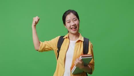 녹색 스크린 배경 스튜디오에 서 있는 동안 미소 짓고 팔다리를 구부리는 배과 몇 권의 책을 가진 아시아 여성 학생