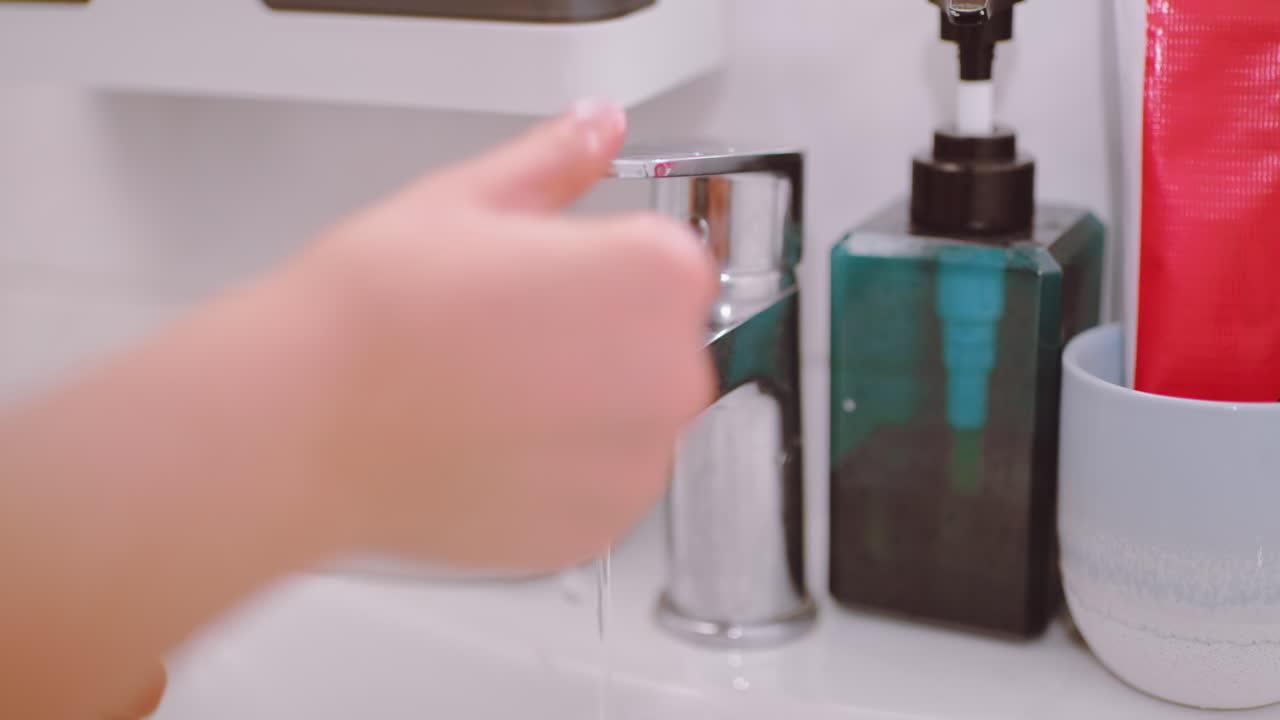 Close up of hand opening faucet in bathroom sink letting water flow before turning it off, showing daily hygiene action with soap, dispenser, toothbrush and toothpaste nearby