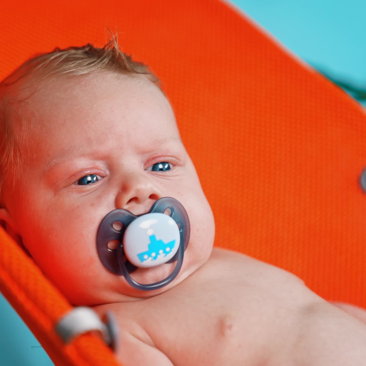 Unhappy blue-eyed blond Caucasian infant with a pacifier in mouth. Portrait of a kid in orange chair. Close up