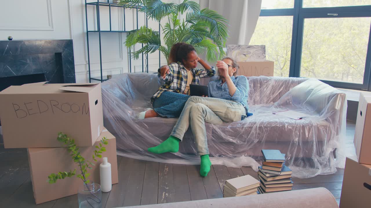 Diverse couple moving into spacious apartment and discussing sits on sofa