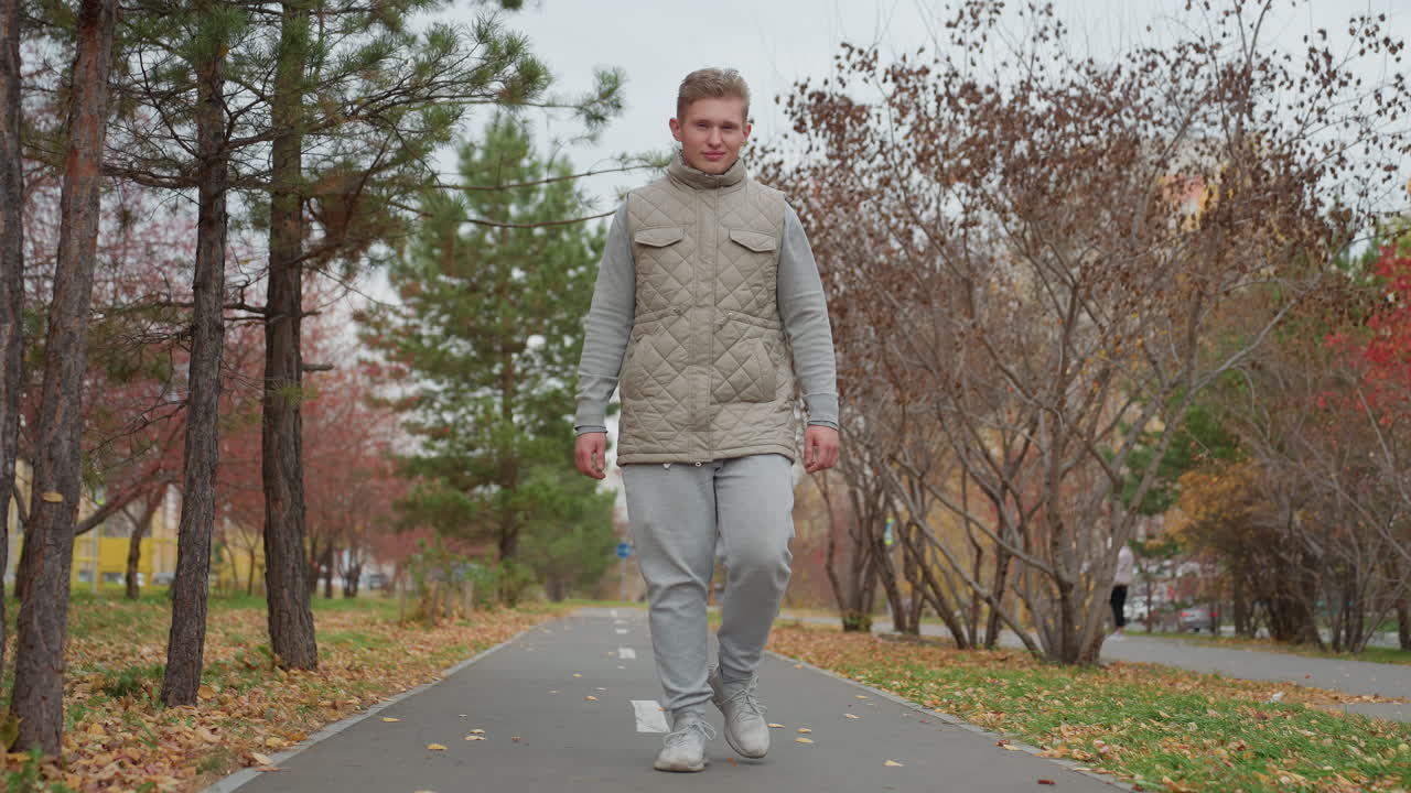 Young strong man walking along tarred road in park surrounded by autumn trees with dry leaves while people walk in background on overcast day in peaceful outdoor setting during fall season