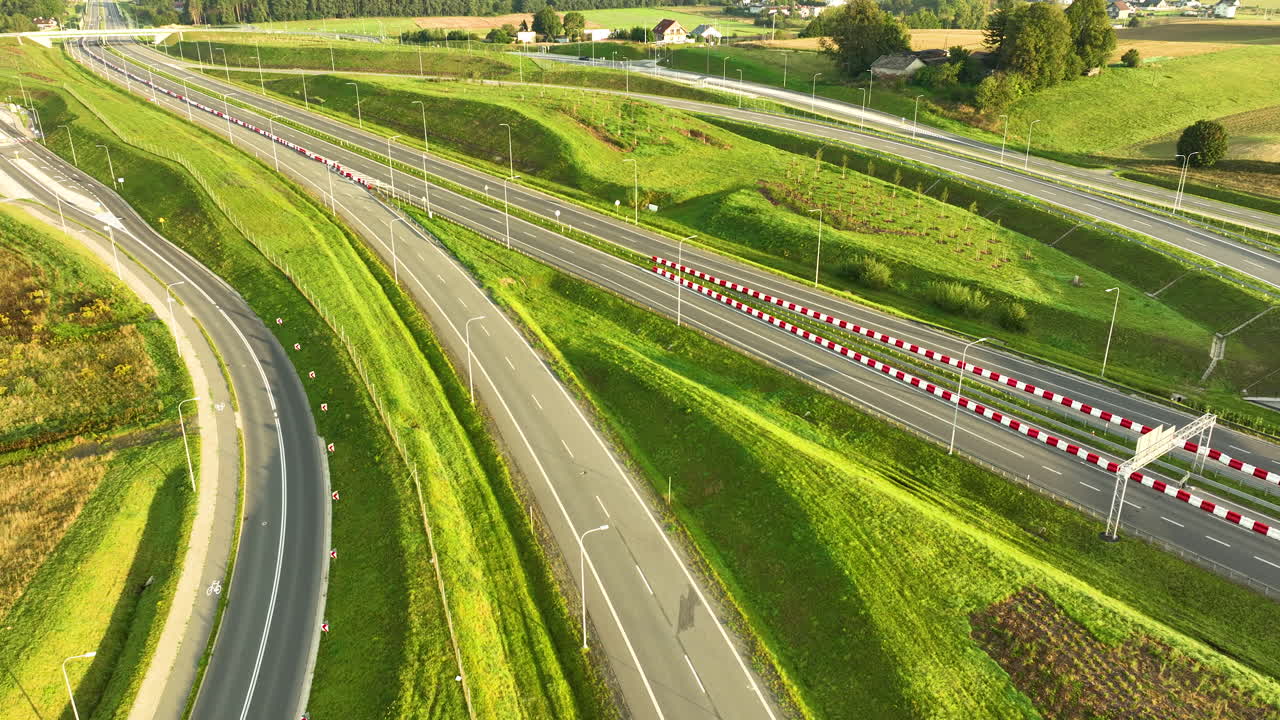 Sunlight hits a modern, sweeping highway junction surrounded by vibrant green, sculpted slopes. Two cars drive past red-and-white safety barriers in this busy aerial shot
