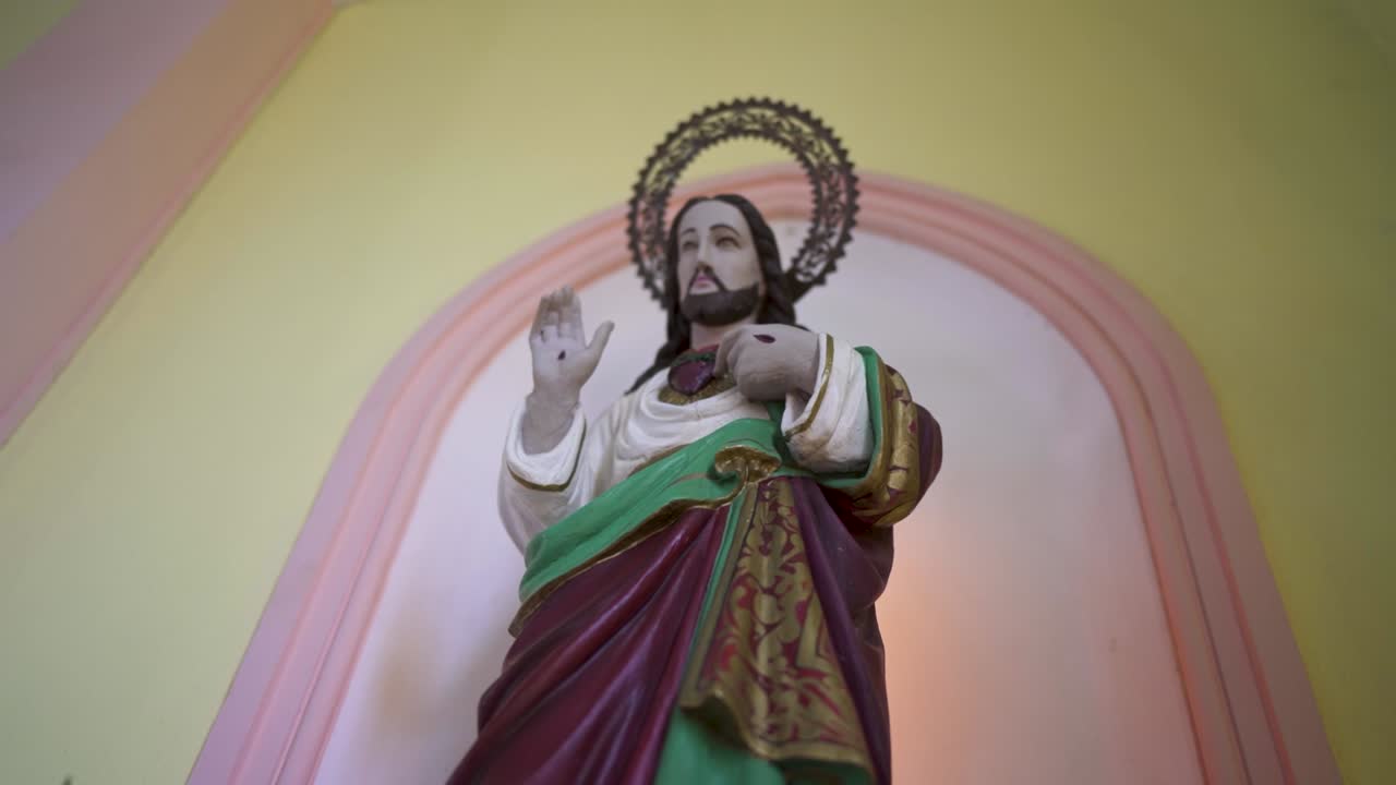 Jesus Christ stands inside a church niche, with maroon robes and gold detailing, one hand raised in blessing, other near the chest, framed by metal halo and soft pink arch against a yellow background
