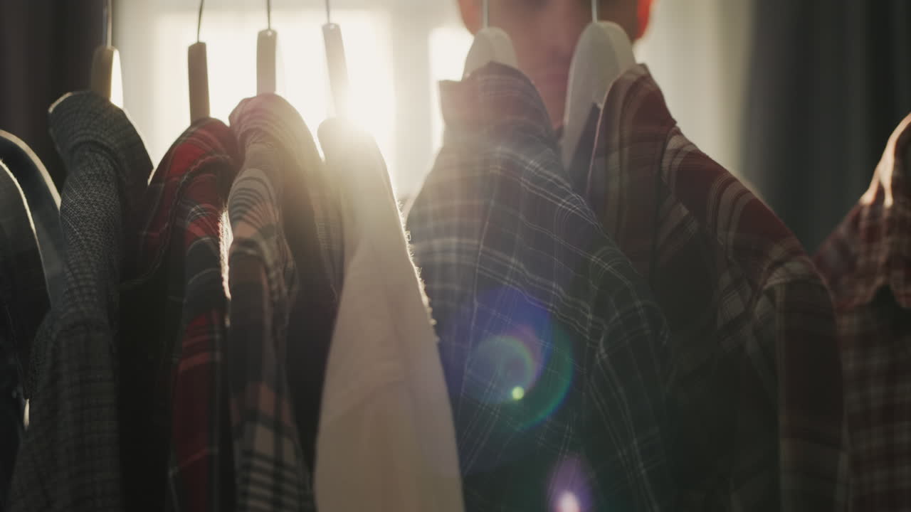 A young man chooses a shirt in a dressing room or shop. The sun from the window illuminates the rack with hangers