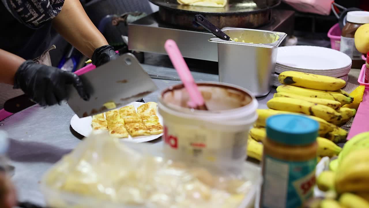 A vendor prepares roti with bananas and toppings at a bustling street market in Phuket, Thailand. Bright, colorful, and lively atmosphere
