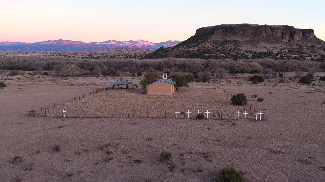 Cinematic drone orbit of a rural church at dusk reveals white crosses on a perimeter fence. The vast, arid landscape features a massive mesa and snow-capped peaks. Ideal for documentary openings