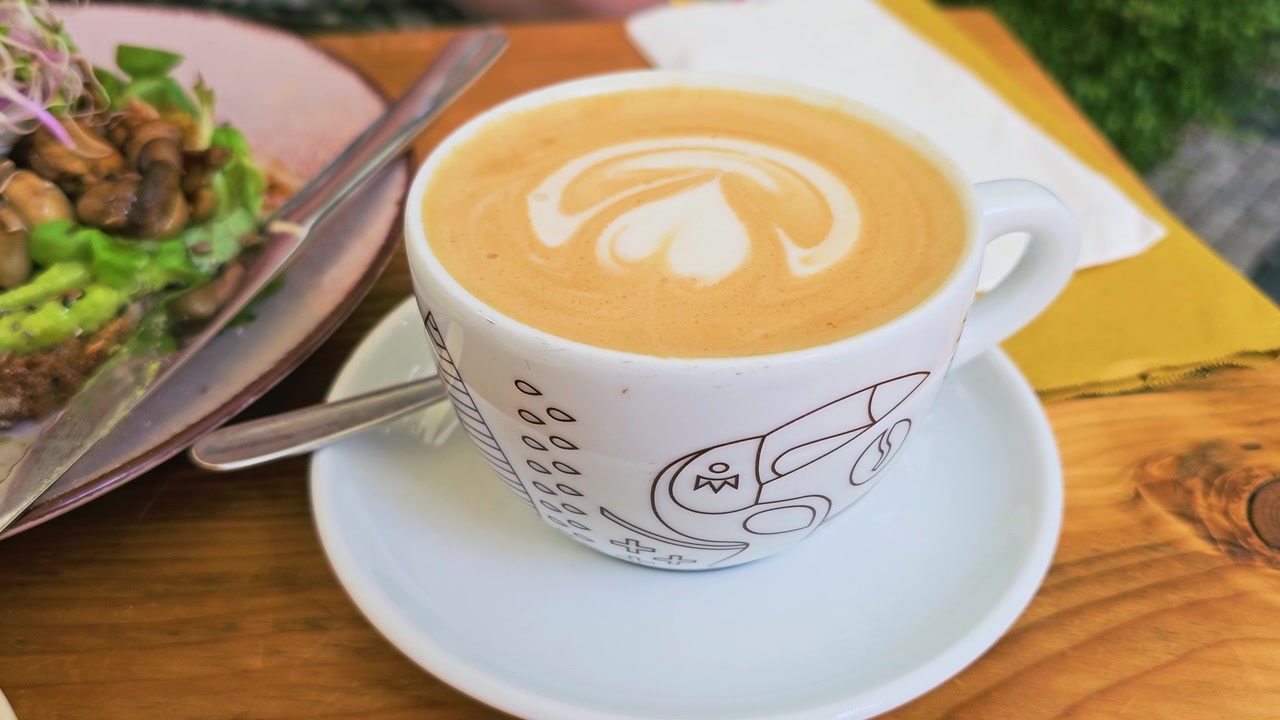 Caffè cappuccino fresco con latte art in schiuma servito in una tazza di ceramica bianca, ripreso da vicino su un tavolo di legno di un ristorante con toast gourmet all'avocado sullo sfondo a colazione o brunch.