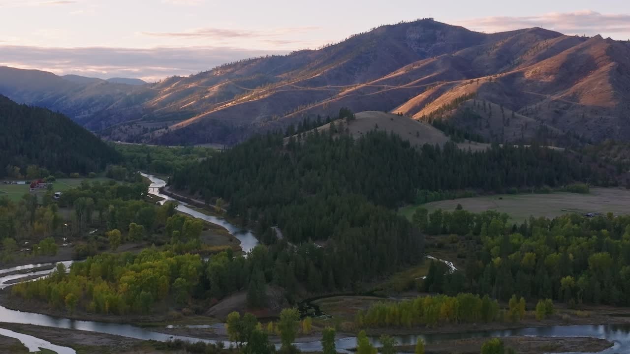 Golden sunset light illuminating rugged mountain slopes and forested hills above winding river valley, aerial drone, tracking left