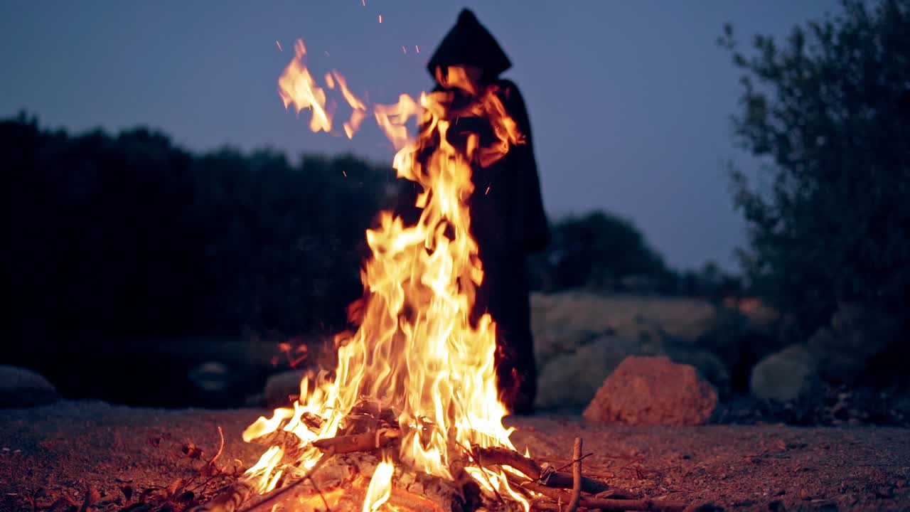 Horrible black silhouette behind the bright flame outdoors. Vivid flame from fire in the evening and witch in black costume waving arms making fear.