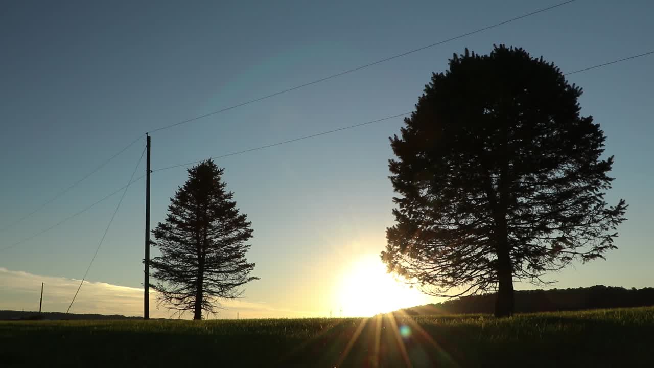 A lone car drives by on rural country road at sunset - silhouetted powerlines and trees - establishing shot