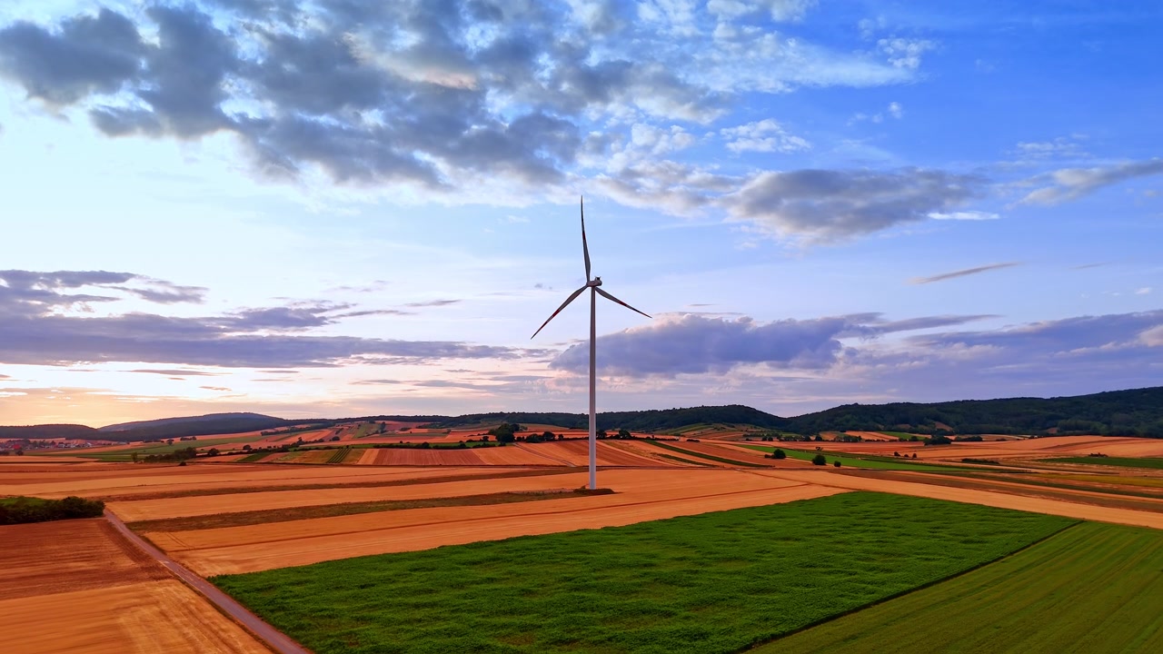 Wind turbine operating in rural fields before distant hills. Drone shot of a single green energy generator surrounded by yellow crops under blue sky with white clouds.