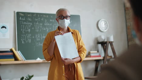 Female-Teacher-in-Mask-Talking-with-Student-on-Lesson