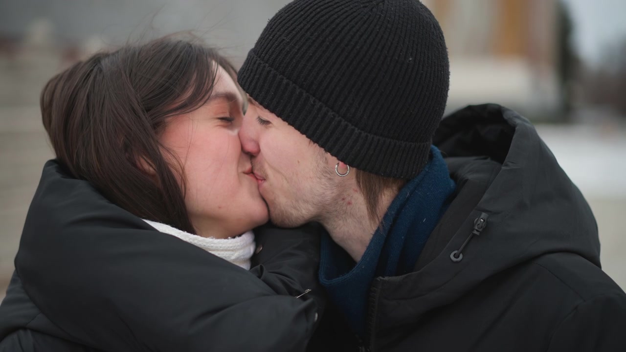 Close up of young woman and man outdoors in cold weather progressing from looking away to smiling together. scene shows them wearing winter clothing with soft lighting against urban