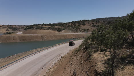 Drone-Siguiendo-A-Un-Camionero-Rojo-Conduciendo-Lentamente-Por-Una-Carretera-En-Turquía-Karaman-Entre-El-Lago-Azul-Y-Las-Montañas-En-Un-Verano-Soleado-Aunque-Con-Curvas