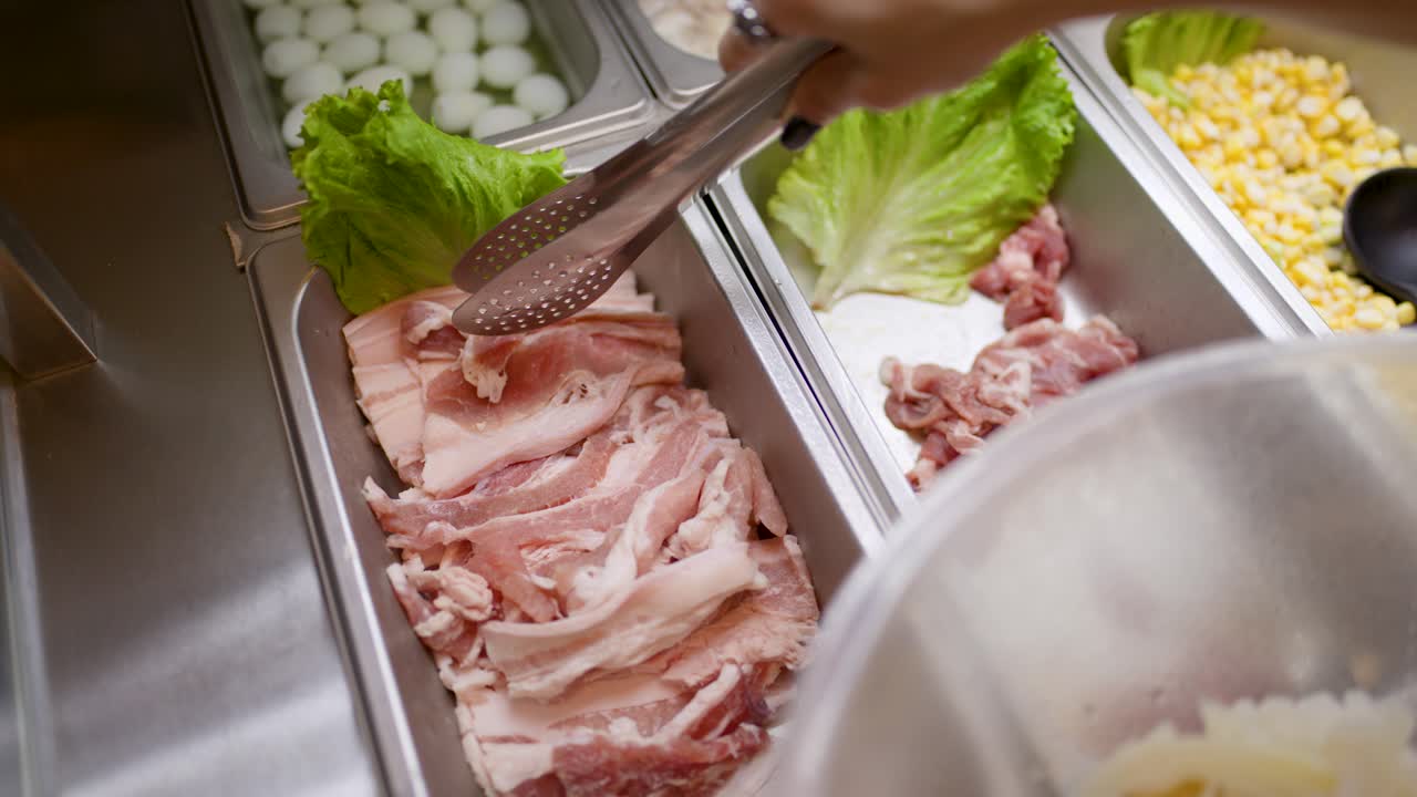 Hand uses tongs to pick sliced raw meat from buffet tray under bright kitchen lighting