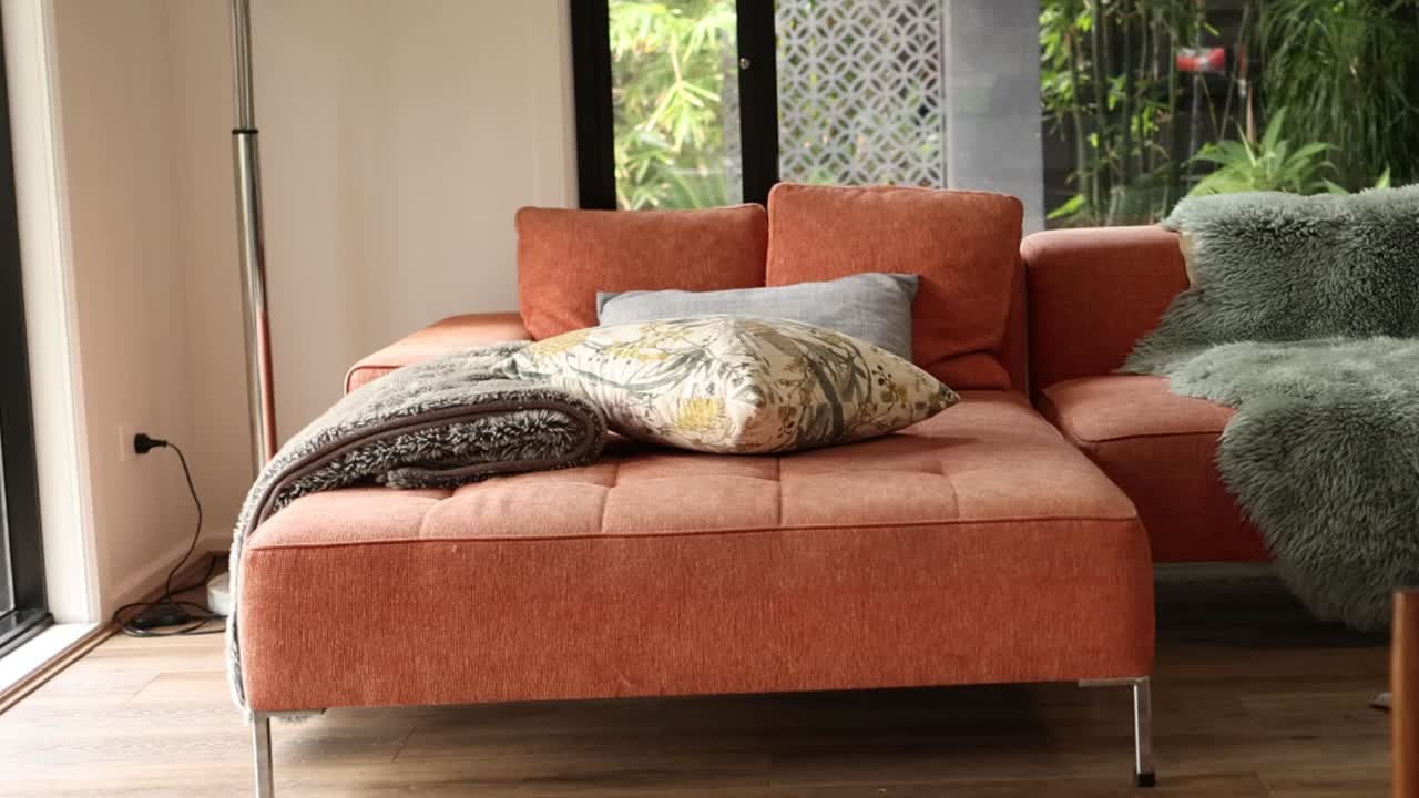 A contemporary orange sofa features plush cushions and throws against a lush garden backdrop