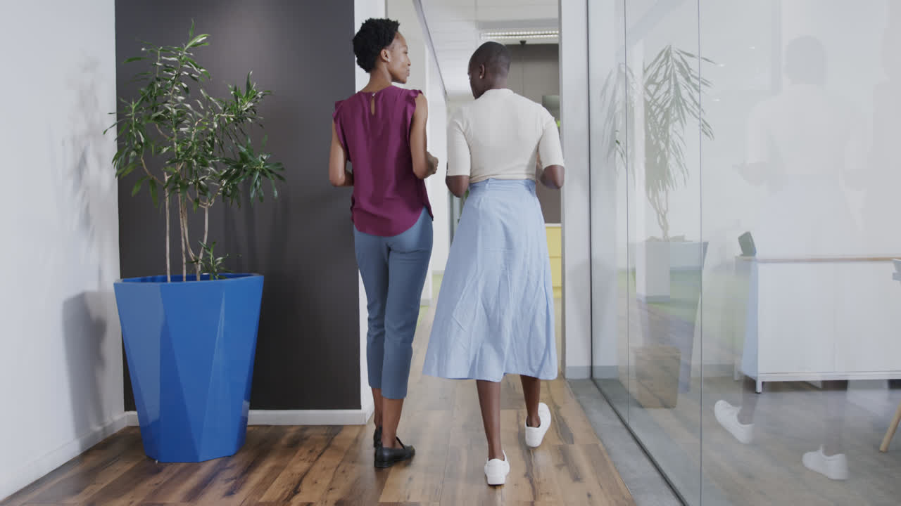Rear view of two african american businesswomen talking, walking in office corridor, in slow motion