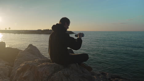 during-golden-hour-man-sits-by-seaside,-he-opens-thermos-pours-drink-enjoys-it,-then-closes-lid-and-leaves-the-scene