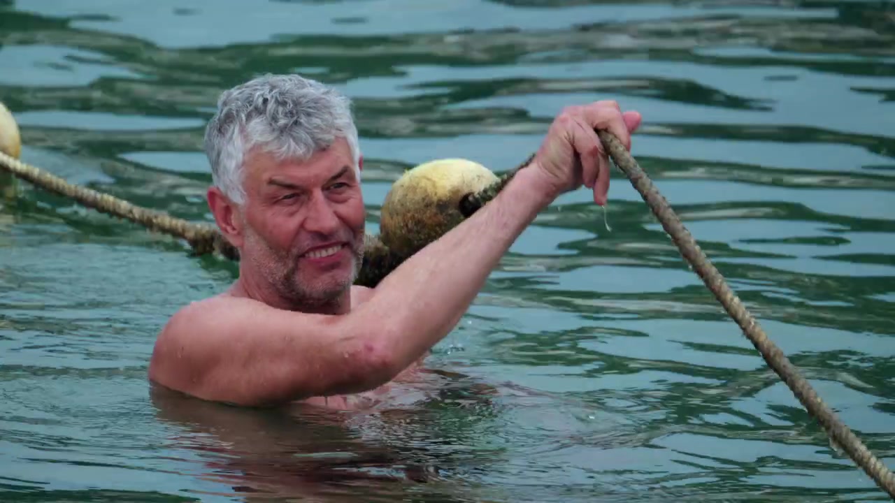 Anziano che nuota in mare, aggrappato a una boa e a una corda, mostrando un'espressione naturale di relax e godendosi il piacere del mare.