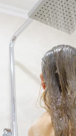 in a bright bathroom, a millennial woman lathers shampoo onto her hair, fully embracing the relaxing experience of showering and self-care amidst wooden blinds.