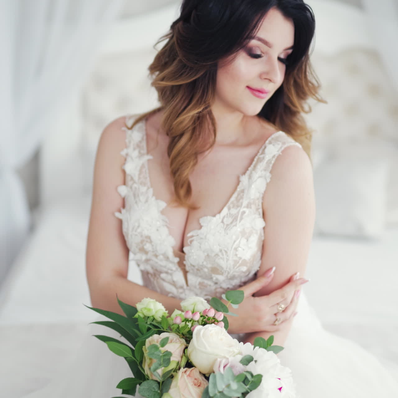 Video shot of a young bride in a wedding dress on the background of a bed. Attractive model with modern hairstyle in white dress is posing to camera inside the light room.