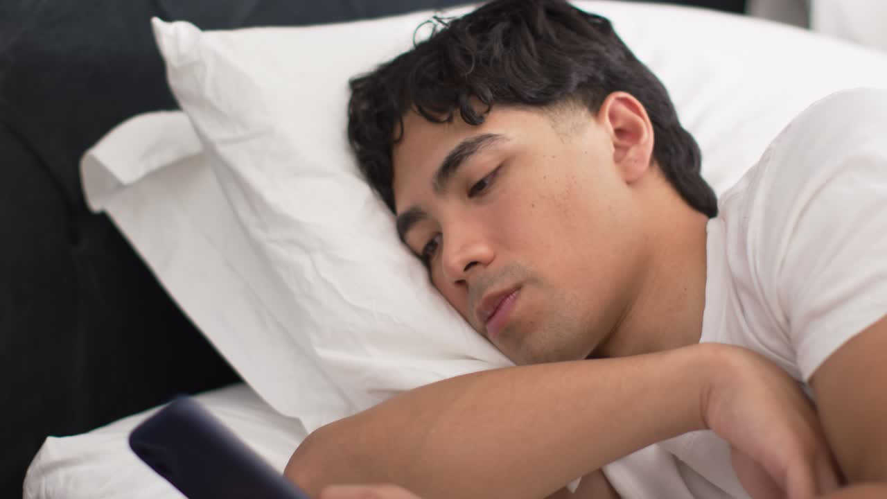 ベッドに横たわるアジア人男性が、目覚まし時計（5時38分）の横でスマートフォンに手を伸ばし、スクロールしている。