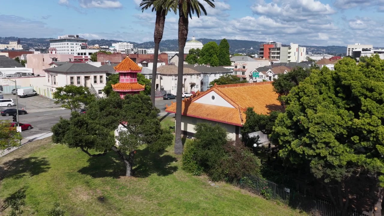 Torre pagoda cinese con tegole arancioni accanto a un'alta palma, parco verde e quartiere urbano misto che si estende verso lo skyline del centro e le colline sotto un cielo soleggiato. California, USA. Orbita aerea.