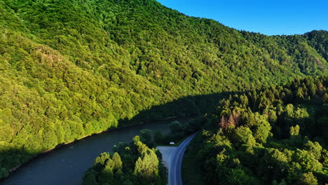 narrow river flows at the foot of the mountains covered with lush woods. drone footage over the highway along the river. slovakia