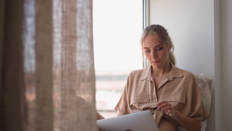 Woman-writes-report-via-computer-sitting-on-sill-at-home