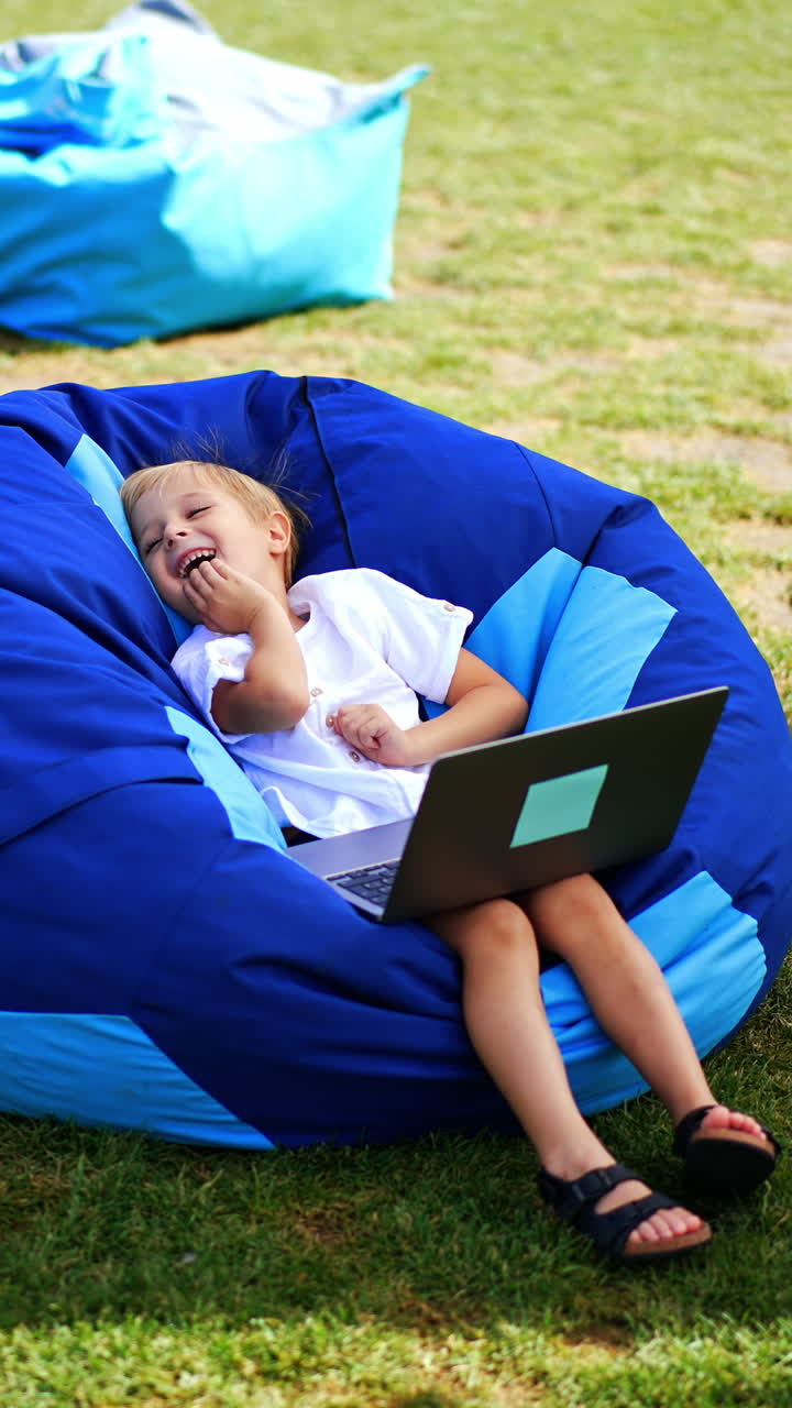 Cute boy in white shirt sits in bean bag chair with laptop on his knees. Kid watched video and laughs leaning back in chair. Vertical video