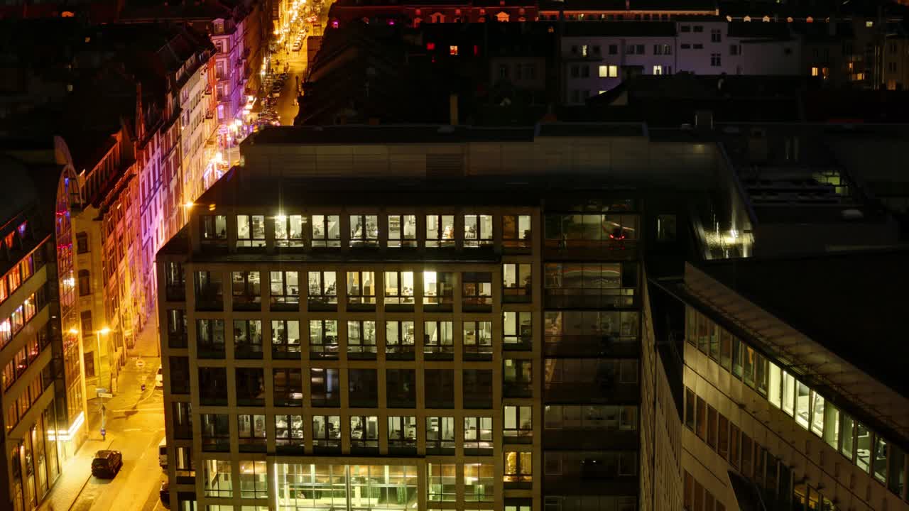 este timelapse presenta un vibrante paisaje nocturno urbano visto desde un ángulo alto, mostrando edificios modernos con numerosas ventanas iluminadas, y farolas naranjas que bordean las calles