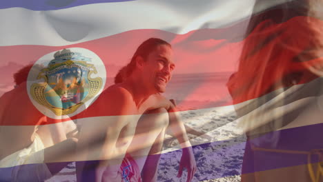 Animation-of-flag-of-costa-rica-waving-over-diverse-friends-sitting-and-enjoying-at-beach