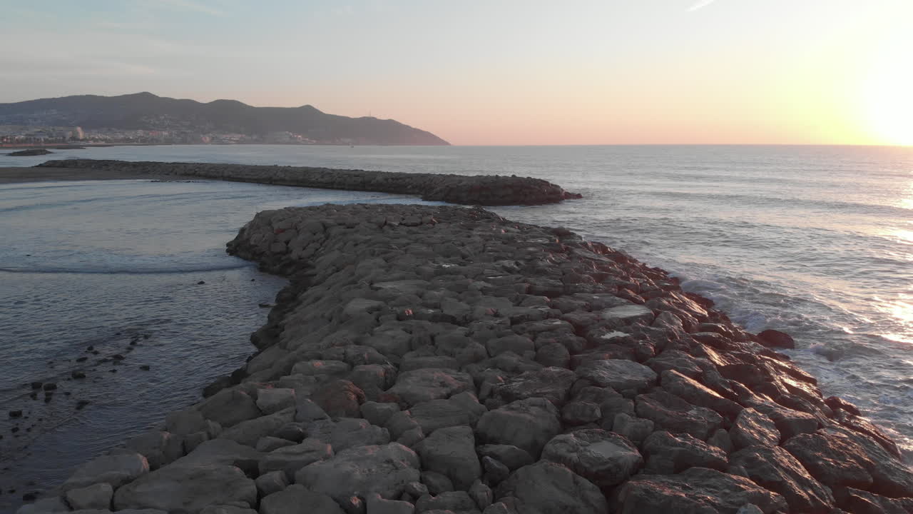 잔잔한 파도에 씻겨진 석조 부두 위를 드론으로 저공비행하며 아침 햇살이 비치는 바다로 뻗어 해안과 산의 탁 트인 전망을 감상할 수 있습니다.