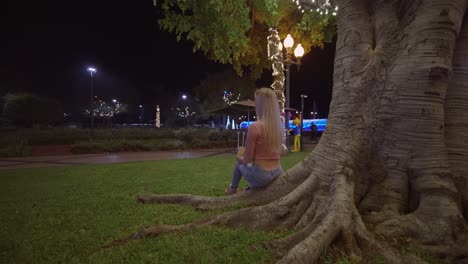 Detrás-De-Una-Mujer-Rubia-Sentada-En-La-Raíz-Del-árbol-En-El-Parque-Por-La-Noche-Comiendo-Castañas,-Funchal