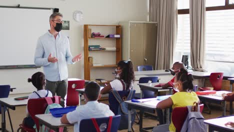 Caucasian-male-teacher-wearing-face-mask-teaching-students-in-the-class-at-school