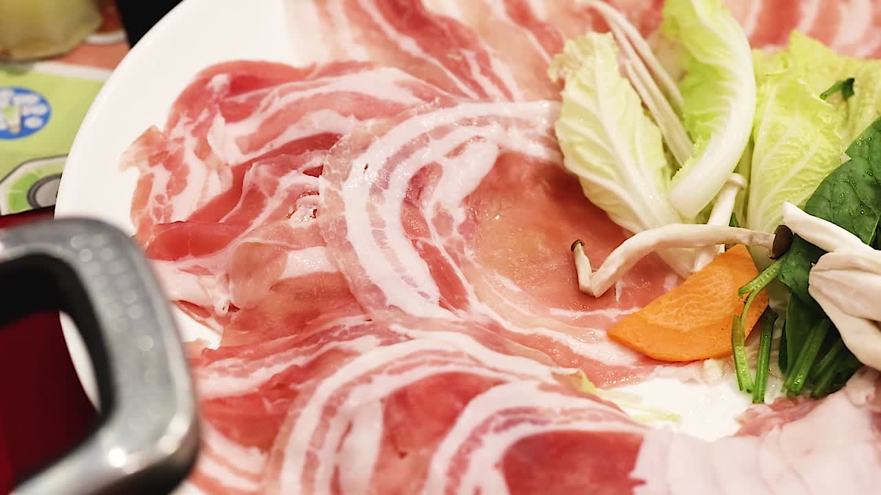 Close-up of pork slices and vegetables being arranged with chopsticks on a plate for hotpot preparation.