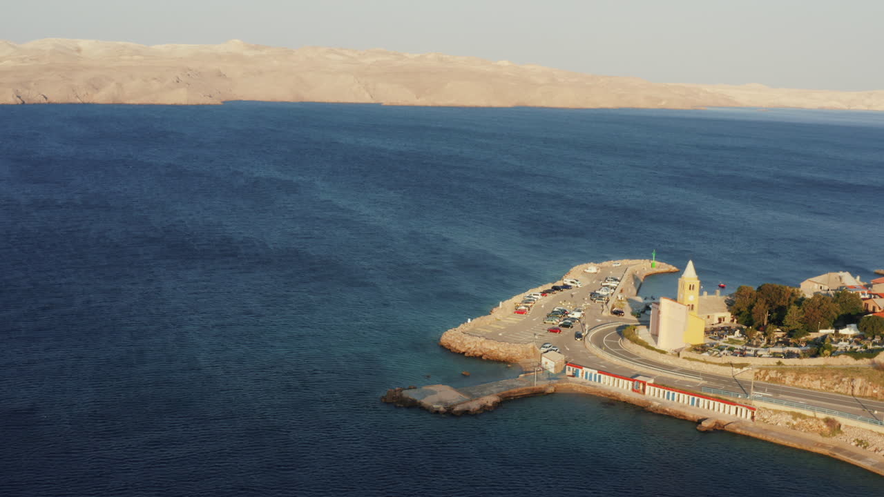 Aerial panorama of dark blue textured sea, scenic peninsula with church and harbor and distant coastline on the horizon. Sunrise on a sunny day. Pag Island, Croatia, Adriatic