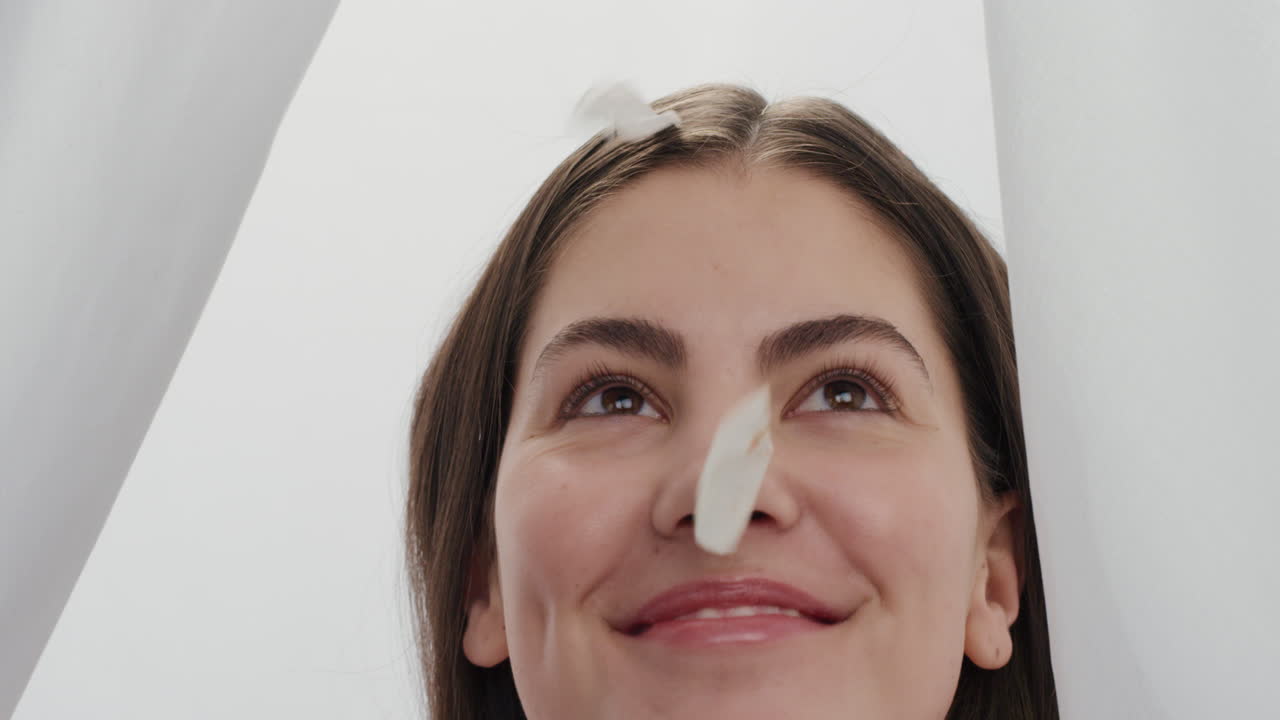 retrato en cámara lenta hermosa joven pétalos de rosa cayendo sobre atractiva mujer sonriendo disfrutando del lujo entre las sábanas en fondo blanco concepto de belleza