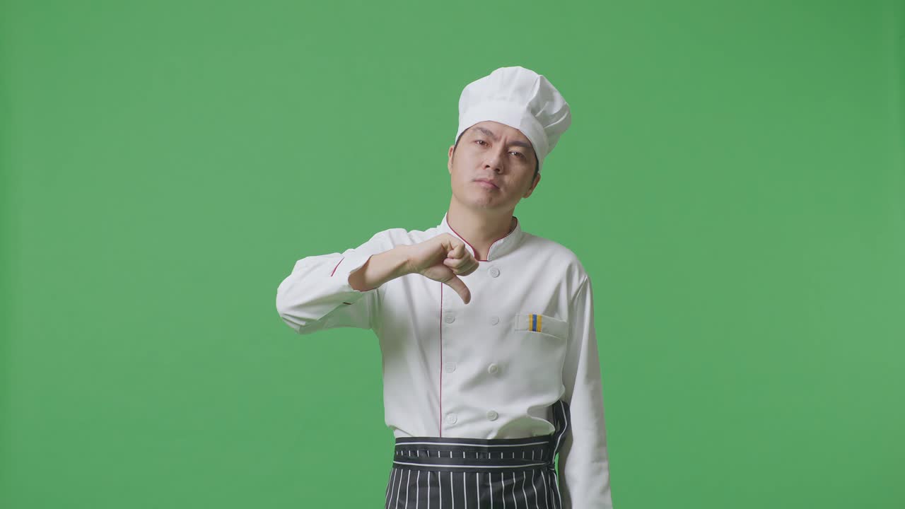 el chef asiático muestra los pulgares hacia abajo en un gesto a la cámara mientras está de pie en el estudio de fondo de la pantalla verde