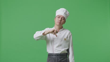 el chef asiático muestra los pulgares hacia abajo en un gesto a la cámara mientras está de pie en el estudio de fondo de la pantalla verde