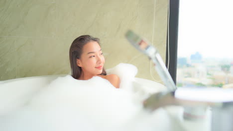 beautiful asian woman in a bathtub full of foam looking through window, spa day relaxation treatment, full frame