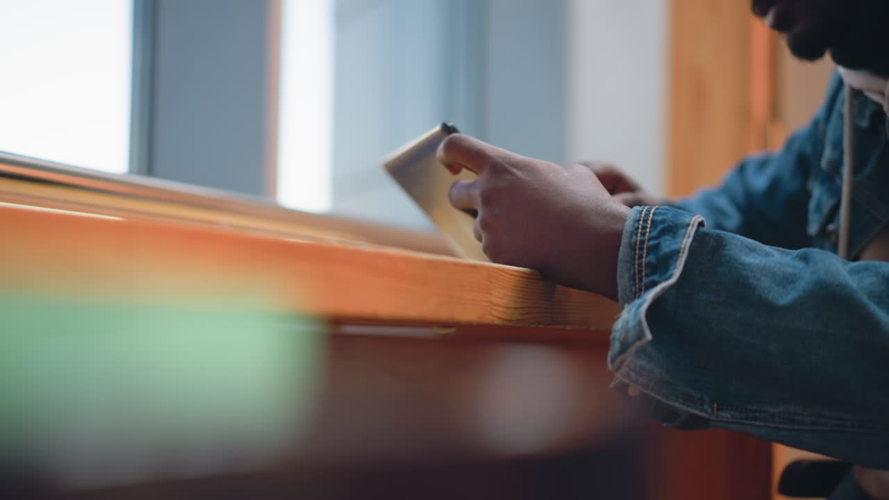 Close-up of person holding smartphone on wooden windowsill table, denim jacket sleeve visible, natural light streaming through window, casual workspace ambiance, hands scrolling touchscreen