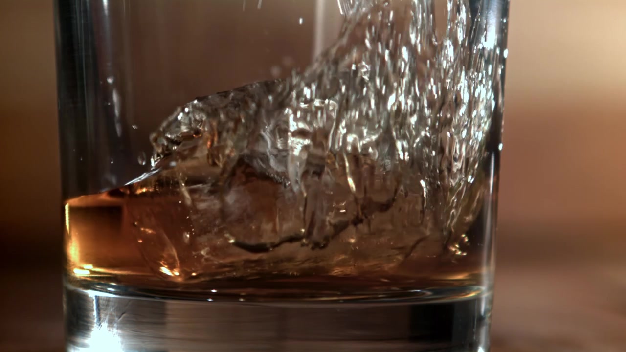 Cubo di ghiaccio che cade in un bicchiere di whisky con liquido versato, al rallentatore