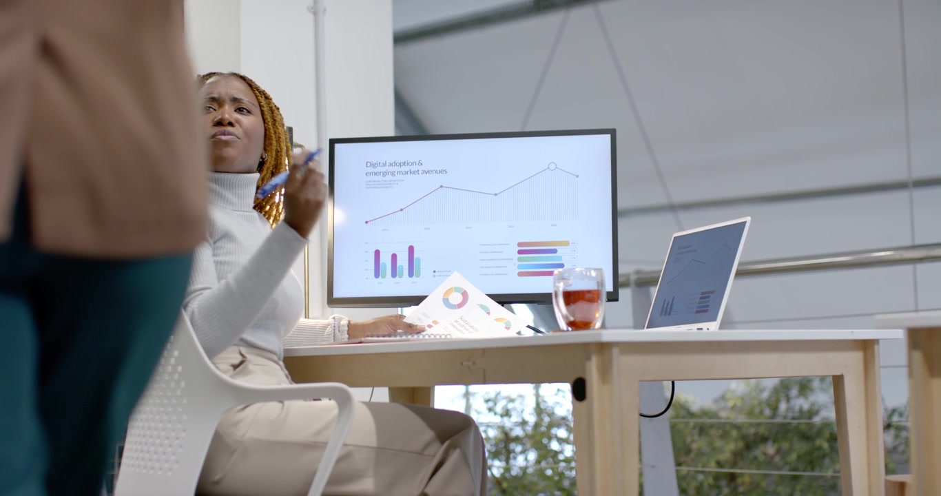 Diverse team African woman reading report on screen at desk pointing and speaking as peer passing