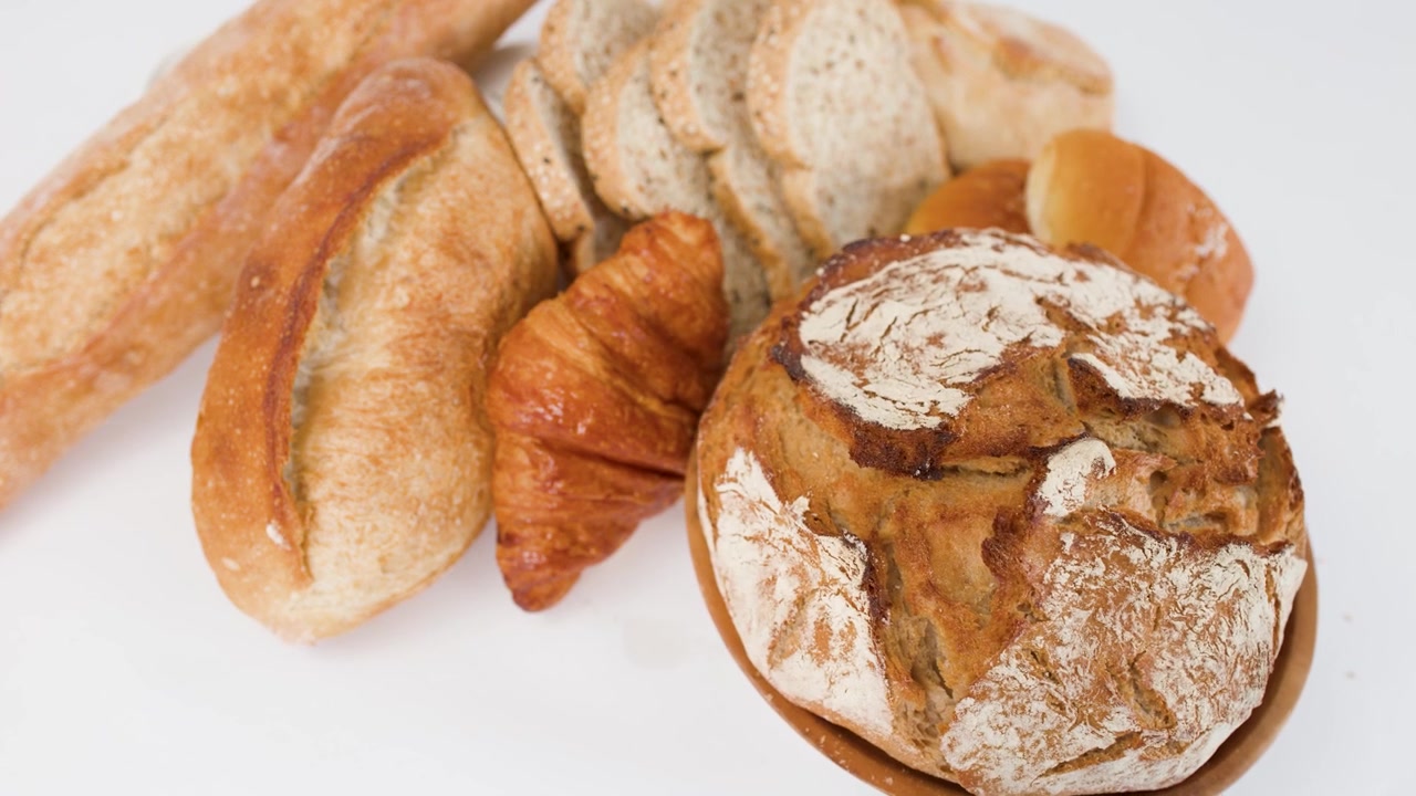 Ripresa a rotazione lenta di una pagnotta di pane a lievitazione naturale, un croissant e una baguette su una superficie bianca pulita.