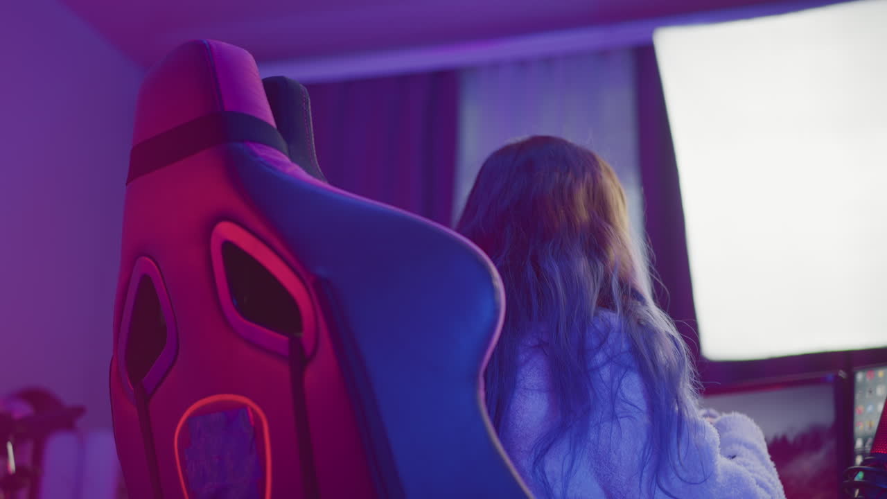 rear view of woman in fluffy jacket seated in gaming chair picking up white headphones while colorful led light panel glows behind dual monitors and red microphone stands next to rgb keyboard on desk