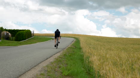 professional road cyclist man cycling uphill on a winding country road by crop field low angle rear view