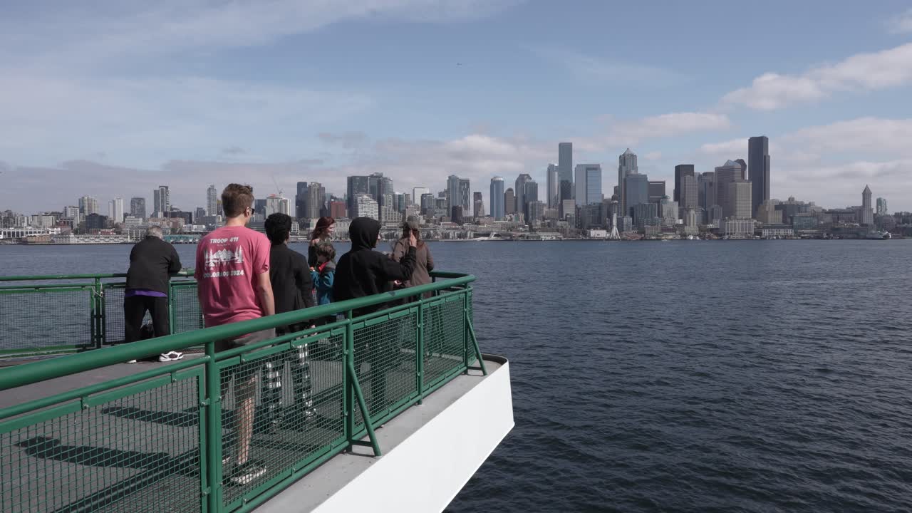 Group of tourists enjoying the view of Seattle's skyline, including the Space Needle, from the deck of a ferry on a sunny day.